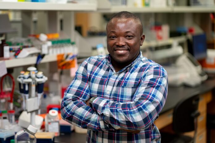 Smiling man with arms crossed in a laboratory, wearing a blue plaid shirt.